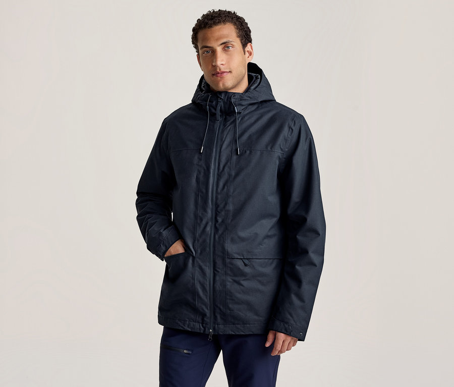 Ein Mann in einer dunkelblauen Kapuzenjacke und dunkelblauen Hosen steht mit einer Hand in der Jackentasche und blickt nach vorne.
