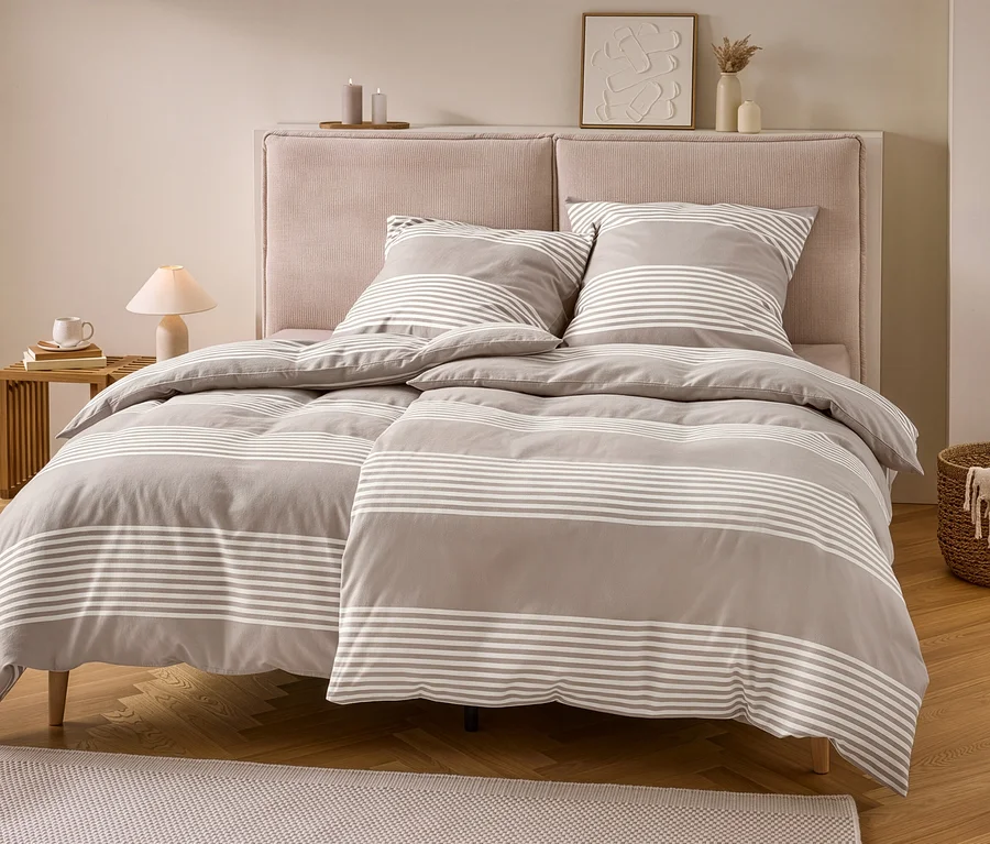 Ein Bett mit grauer Biber-Bettwäsche mit weißen Streifen, bestehend aus zwei Kissen und zwei Decken. Dazu ein rosa Kopfteil, ein Nachttisch mit Lampe und ein Korb mit Decke.