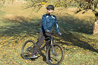 Ein bärtiger Mountainbiker mit Funktions-Jogginghose, Shirt, Helm und Sonnenbrille steht an einem sonnigen Tag an einem Hang und geniesst seine Tour.