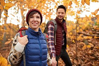 Steppwesten zum Wandern: Eine junge Frau, die eine rote Mütze und eine blaue Sportweste trägt, hält ihren Freund an der Hand und geht im herbstlichen Wald spazieren.