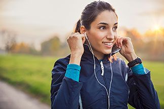 Regenjacken für den Sport: Eine junge, dunkelhaarige Frau, bekleidet mit einer blauen Softshelljacke und weissen Kopfhörern im Ohr, macht sich bereit zum Joggen in der Natur.