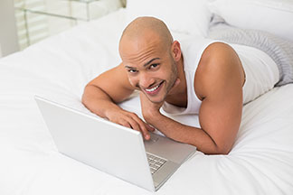 Ein Mann, der ein weisses Unterhemd und eine lange Pyjamahose trägt, liegt im Bett und arbeitet an seinem Laptop.