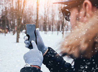 Handschuhe für Herren mit wichtigen Details: Ein Mann steht in einer verschneiten Winterlandschaft und bedient mit seinen Handschuhen an den Händen sein Smartphone.