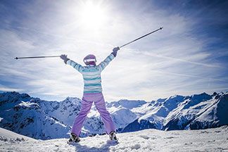 Eine Frau auf Skiern reckt die Arme mit Skistöcken in die Höhe und schaut auf ein Bergpanorama.
