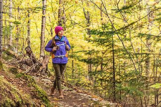 Eine junge Frau, die eine pinke Mütze, einen Wanderrucksack und eine lila Softshelljacke trägt, wandert durch einen herbstlichen Wald.