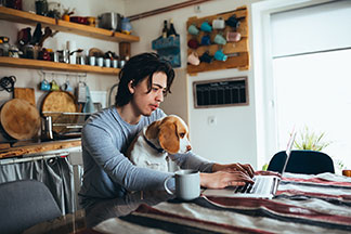 Ein junger Mann, der Homewear trägt, arbeitet an seinem Küchentisch an seinem Laptop und hat seinen Hund auf dem Schoss.