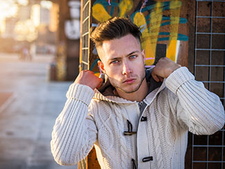 Ein junger Mann, der eine weisse rustikale Strickjacke mit Kapuze trägt, steht vor einer Wand, die mit Graffiti besprüht ist.