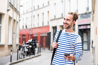 Ein fröhlicher, junger Mann in einem blau-weiss geringelten Langarmshirt spaziert mit einem Smartphone in der Hand durch eine Stadt.