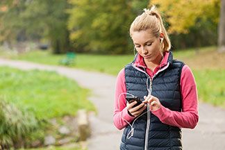 Eine junge, blonde Frau, die eine blaue Sportweste trägt, geht durch einen Park und schaut dabei auf ihr Handy – dabei hat sie Stöpsel in den Ohren und hört über das Telefon Musik.