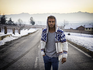 Ein junger Mann, der eine weisse Strickjacke mit Norwegermuster und Kapuze trägt, läuft bei Sonnenaufgang auf einer winterlichen Strasse durch eine nebelverhangene Landschaft.
