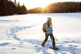 Eine junge Frau, die eine dicke, blaue Winterjacke, eine schwarze Softshellhose und eine weisse Mütze trägt, läuft mit Schneeschuhen und Stöcken durch die verschneite Winterlandschaft.