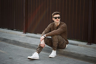 Sportschuh für den Alltag: Ein stylisch gekleideter, junger Mann, der weisse, knöchelhohe Sneaker trägt, sitzt lässig an einer Bordsteinkante.
