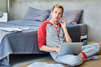 Ein junger Mann sitzt telefonierend auf dem Fussboden an sein Bett gelehnt, arbeitet an seinem Laptop und ist dabei mit einem Homewear-Set bekleidet.