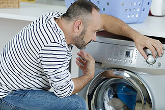 Ein Mann, der eine Jeans und einen weiss-schwarz gestreiften Pullover trägt, kniet vor der Waschmaschine, um seine Jogginghose zu waschen, und stellt das Programm ein.