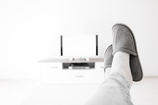 Herrenschuhe für daheim: Nahaufnahme von den Beinen eines Mannes, die ausgestreckt auf einem Tisch abgelegt sind. An den Füssen trägt der Herr Pantoffeln.