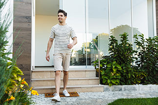 Ein junger Mann in einem schwarz-weiss gestreiften Oberteil und beigen Chino-Shorts verlässt beschwingt sein Haus.