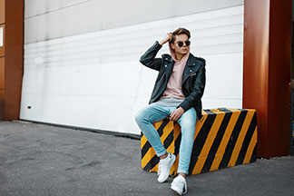 Herren Halbschuhe für den Sommer: Ein junger Mann in Jeans, Kapuzenpullover und weissen Sneakers sitzt lässig auf einem schwarz-gelb gestreiften Betonblock vor einer Garage.