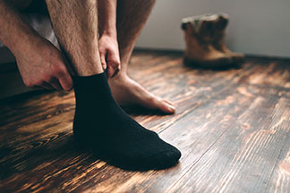 Nahaufnahme der Beine eines Mannes, der auf seinem Bett sitzt und sich seine Strümpfe anzieht. Ein Fuss trägt schon einen schwarzen Socken, der zweite ist noch barfuss.