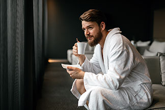Ein junger Mann in einem weissen Bademantel trinkt, auf der Bettkante sitzend, einen Tee aus einer weissen Tasse.