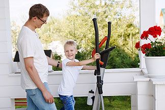 Fitness für zuhause: Ein kleiner Junge mit seinem Vater auf einem Crosstrainer auf der heimischen Terrasse.