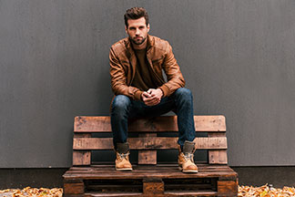 Top-Qualität für Ihre Herrenschuhe: Ein Mann, der braune Boots und eine braune Lederjacke trägt, sitzt draussen auf einer Bank aus Holzpaletten.