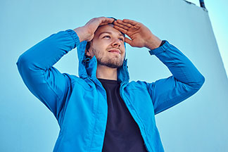 Übergangsjacke für draussen: Ein junger Mann, der eine blaue Softshelljacke trägt, hält sich die Hände an die Stirn und schaut in die Ferne.