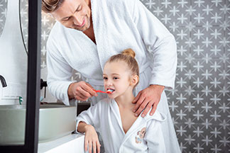 Ein Vater und Tochter, die beide jeweils einen weissen Bademantel tragen, stehen im Badezimmer und der Vater putzt der Tochter die Zähne