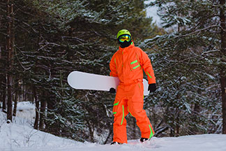 Ein Snowboarder der einen orangenen Skianzug trägt, läuft abseits der Piste durch einen verschneiten Winterwald.