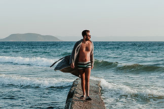 Ein junger Mann steht am Strand und fährt sich mit der Hand durchs Haar. Er trägt eine grün-blau gestreifte Herren-Badehose und eine Sonnenbrille.