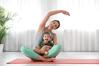 Yoga-Kleidung in Top-Qualität: Eine junge Mutter sitzt auf ihrer Yogamatte und hebt den Arm über den Kopf, Ihr Sohn sitzt auf ihrem Schoss.