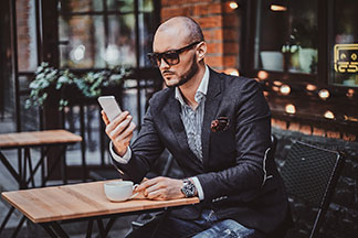 Qualität für Ihre Herrenuhr: Ein junger Mann im Anzug sitzt im Aussenbereich eines Cafés und schaut auf sein Handy. Am Arm trägt er eine Herrenuhr.