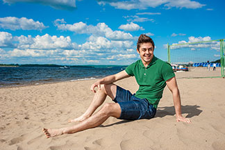 Ein junger Mann in weiten Herren-Badehosen sitzt gemütlich im Schatten an einem tropischen Strand.
