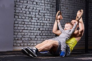 Ein junger Vater in Sportshorts hält seinen Sohn auf dem Schoss, und trainiert gleichzeitig seine Arme an Ringen im Fitnessstudio.