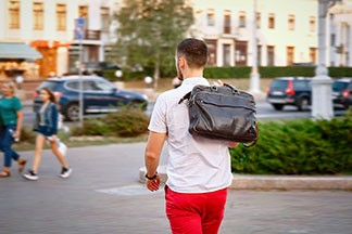 Ein junger Mann in roten Hosen und einem weissen Kurzarm-Hemd geht mit seiner Leder-Aktentasche durch die Stadt, die er über die Schulter geworfen hat.