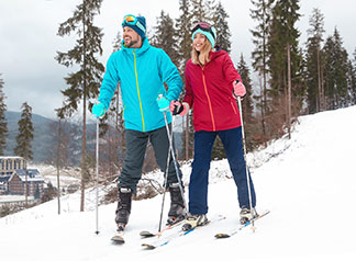 Die richtige Skibekleidung für die Piste: Ein junges Pärchen, das jeweils ein zweiteiliges Skioutfit trägt, steht zufrieden und glücklich lächelnd auf einer Skipiste.