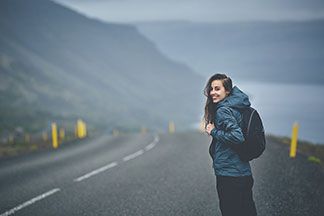 Gut ausgerüstet: Eine junge Frau, die eine Regenjacke und einen Rucksack trägt, läuft an einem regnerischen Tag auf einer verlassenen Strasse durch die Natur.
