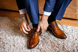 Herrenschuhe fürs Büro: Nahaufnahme der Füsse eines Mannes, der einen blauen Anzug trägt, auf seinem Bett sitzt und sich seine braunen Lederschuhe bindet.