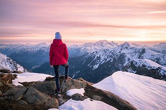 Ein junge Frau in einer roten Softshelljacke steht in einer winterlichen Gebirgslandschaft und schaut in die Ferne in Richtung verschneiter Berge.