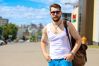 Ein junger Mann, der ein weisses Herrenunterhemd, eine Sonnenbrille und einen Rucksack trägt, schlendert lässig durch eine Stadt.