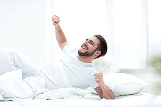 Ein junger Mann, der ein weisses T-Shirt trägt, liegt am Morgen noch halb unter seiner weissen Decke und streckt sich zufrieden in seinem Bett.