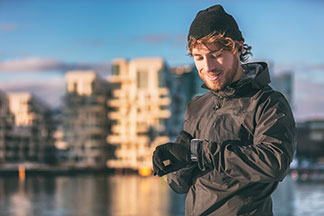 Handschuhe für den Sport: Ein junger Mann, der gerade joggen gehen möchte, eine schwarze Mütze, Sportkleidung sowie Handschuhe für Herren aus Fleecematerial trägt, schaut auf seine Uhr.