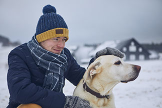 Das ideale Accessoire für den Winter: Ein junger Mann, der Schal, Mütze und Handschuhen trägt, streichelt in der Hocke seinen weissen Labrador in einer verschneiten Winterlandschaft.