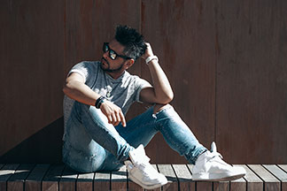 Herrenschuhe für die Freizeit: Ein junger Mann, der weisse Turnschuhe, Jeans, ein graues Shirt und eine Sonnenbrille trägt, sitzt vor einer braunen Wand.