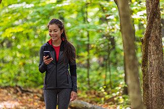 Softshelljacke für den Sport: Eine junge, lachende Frau, die Sportkleidung trägt, läuft durch den Wald und schaut dabei auf ihr Smartphone.
