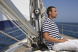 Ein Mann in einem blau-weiss gestreiftem Poloshirt sitzt auf einer Segelyacht und lehnt sich an den Mast.