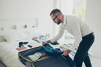 Ein Mann, der ein weisses Hemd und eine schwarze Hose trägt, packt seinen Koffer, der offen auf dem Bett liegt.