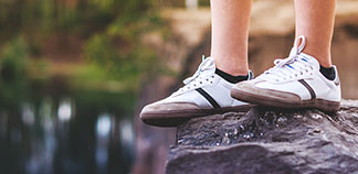 Sportliche Herrenstrümpfe: Nahaufnahme der Beine eines jungen Mannes, der weisse Sneakers und schwarze Sneakersocken trägt, während er auf einem Felsen steht.