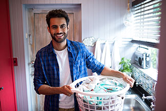 Herrenmode richtig pflegen: Ein junger Mann, der mit einem blauen, offenen Hemd und einem weissen T-Shirt bekleidet ist, trägt einen weissen, runden Plastikkorb voller Wäsche.