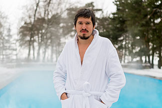 Ein bärtiger Mann in einem weissen Bademantel steht draussen vor einem dampfenden Pool und hält seine Hände in den Bademanteltaschen.