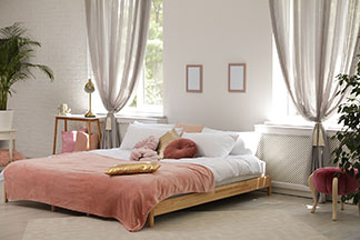 Heimtextilien für Wohlfühlatmosphäre: Ein grosses Schlafzimmer mit einem schön bezogenen Doppelbett mit rosa Tagesdecke, Dekokissen und weissen Vorhängen.